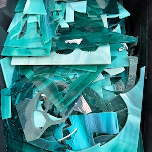 May include: A collection of assorted stained glass scraps in various shades of teal, turquoise, and deep emerald green. The pieces feature smooth, flat surfaces with marbled textures and irregular geometric shapes, all contained within a dark bin.