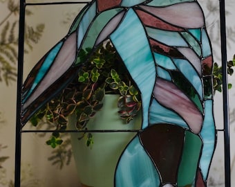 May include: Stained glass panel with a black frame. The design features abstract shapes in shades of blue, teal, brown, and pink. A green pot with a plant is visible behind the glass, adding a natural element to the artwork. The panel is a decorative piece.