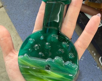 May include: A handcrafted, green glass ornament in the shape of a flask. The glass features a swirling pattern of light and dark green, with bubble-like details. A small metal loop is attached for hanging.