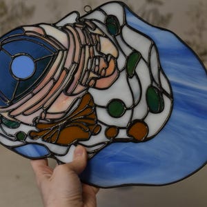 May include: Stained glass art piece featuring a stylized design with a blue, white, orange, and green color palette. The artwork has a dark border and includes a circular blue element. The piece is held up by a hand.