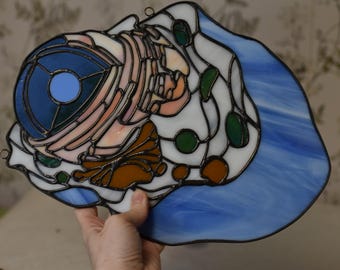 May include: Stained glass art piece featuring a stylized design with a blue, white, orange, and green color palette. The artwork has a dark border and includes a circular blue element. The piece is held up by a hand.