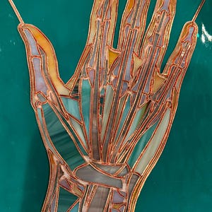 以下が含まれることがあります： 人間の手を描いたステンドグラスのアートワーク。骨と血管が見えます。手は銅で縁取られ、青、緑、グレー、黄色の色合いのカラフルなガラスのセクションがあります。背景は無地のティール色です。