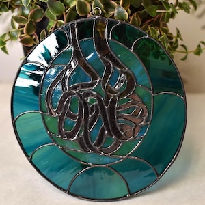 May include: Round stained glass art piece with a teal and green color scheme. The design features a central abstract pattern in dark brown, surrounded by concentric circles of varying shades of teal and green. A small metal loop is at the top for hanging.