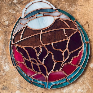 May include: A stained glass art piece with a circular design. The piece features a combination of colors, including blue, brown, red, and purple, with copper-colored lines separating the glass pieces. The artwork is on a tan surface.