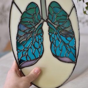 May include: Stained glass artwork depicting lungs in shades of teal and blue, set against a cream-colored background. The oval-shaped piece features a dark metal frame and hanging loops, showcasing a unique anatomical design.