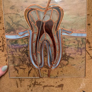 May include: A square stained glass panel featuring a stylised tooth design with a central root canal instrument. The piece uses translucent white, dark brown, and light blue glass segments held together by copper foil lines on a textured background.