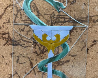 May include: A stained glass panel featuring the Rod of Asclepius symbol. The design consists of a light blue chalice with gold accents and a swirling green glass snake wrapped around the stem and bowl, all set against a clear glass background.