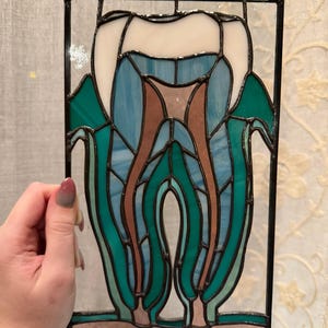 May include: A rectangular stained glass panel featuring a stylized tooth design. The tooth is composed of white, light blue, brown, and teal glass segments held together by black metal solder lines, framed within a thin black metal border.
