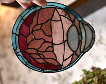 May include: A stained glass piece with a circular shape, featuring a combination of red, pink, and gray glass segments. The design is outlined with black lines and framed by a teal border. The piece is held by a hand.