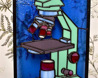 May include: Stained glass art piece featuring a microscope design. The microscope is rendered in shades of green, red, purple, and blue, set against a textured blue background. The piece is framed with a dark border.