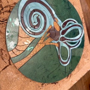 May include: A mosaic artwork featuring a stylized, abstract design in shades of teal, brown, and green. The design includes a spiral and curved shapes, set against a green circular background. The mosaic is set on a brown surface.