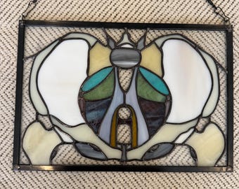 May include: A rectangular stained glass panel featuring an anatomical depiction of a human pelvis. The piece is constructed with white, teal, green, and dark purple glass segments held together by black metal cames and includes a black hanging chain.
