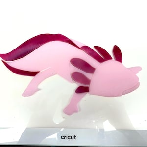 May include: A pink axolotl design with darker pink accents. The axolotl is a stylized, flat design with visible gills and a curved tail. The image is on a white surface, with the word "cricut" visible at the bottom.