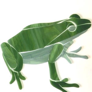 May include: A green frog mosaic with white accent lines. The frog is depicted in a side profile, with its legs extended. The mosaic is made of various shades of green glass, creating a textured appearance. The background is white.