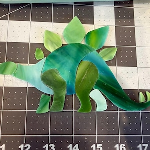 May include: A stained glass stegosaurus in shades of green and blue. The dinosaur has a spiky back and a long tail. The pieces are arranged on a black and white grid.