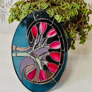 Puede incluir: Obra de arte ovalada de vidrieras con un borde turquesa y un diseño con formas de flores rosas y rojas. La obra de arte se encuentra sobre un fondo de follaje verde.