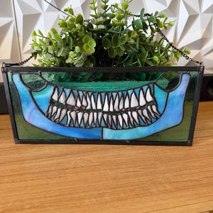 May include: A rectangular stained glass planter featuring a blue and green gradient design with a central graphic of a wide, toothy jaw. The piece is framed in dark metal and includes a black chain for hanging, filled with a green artificial plant.