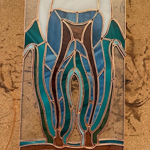 May include: A stained glass panel depicting a human tooth with multiple roots, constructed from various shades of blue, teal, and white glass pieces held together by copper foil solder lines, set against a clear glass rectangular background.