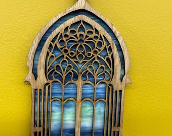 Könnte beinhalten: Holzornament in Form eines Buntglasfensters. Das Ornament hat einen lasergeschnittenen Holzrahmen mit aufwendigen Designs und einen blau-grünen Glaseinsatz. Eine natürliche Schnurschlaufe ist zum Aufhängen befestigt.