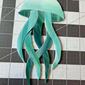 Può includere: Una scultura artigianale di medusa in vetro. La medusa presenta una testa arrotondata azzurra e lunghi tentacoli fluenti in tonalità di verde e verde acqua. Il vetro ha un effetto marmorizzato, che aggiunge profondità e consistenza. Un'opera d'arte decorativa.
