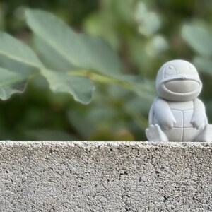 May include: A small, gray, ceramic figurine of a turtle with a friendly expression. The figurine is sitting on a concrete surface with a blurred green background.