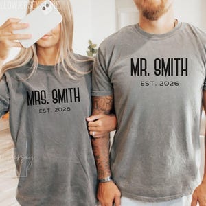 May include: Two gray t-shirts with the text "MRS. SMITH EST. 2026" and "MR. SMITH EST. 2026" printed in black. The shirts are worn by a couple. The shirts are a relaxed fit and appear to be made of a soft material.