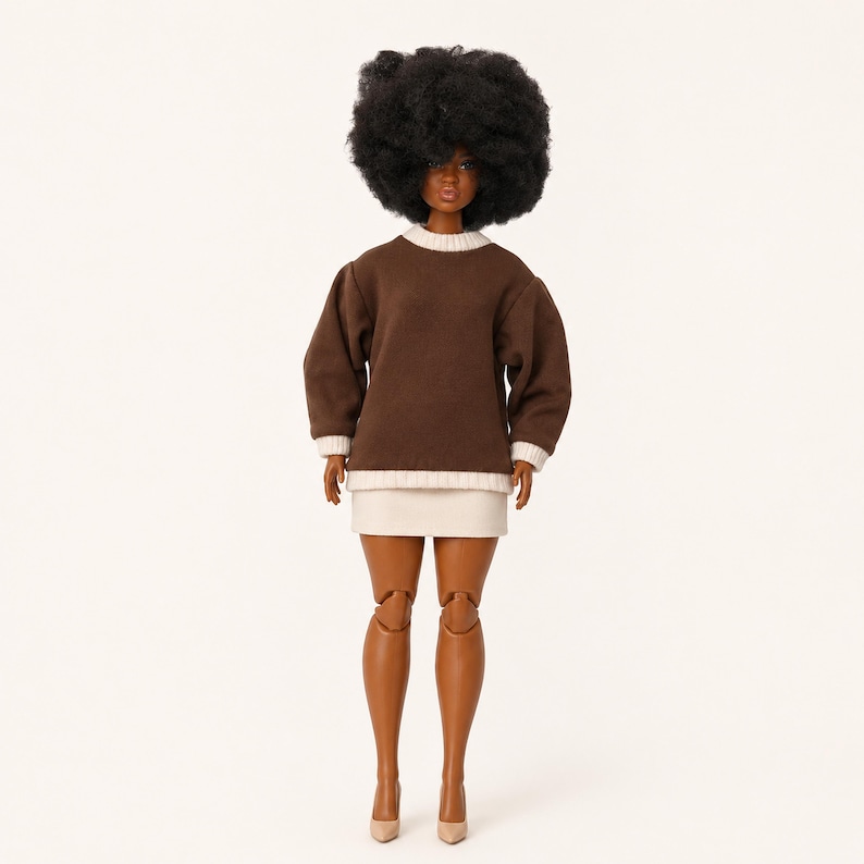 Op de afbeelding: Een pop met een donkere huid en een groot, volumineus zwart afro-kapsel. De pop draagt een donkerbruin sweatshirt met lange mouwen, een cr&egrave;mekleurige geribbelde kraag en zoom, gecombineerd met een korte, cr&egrave;mekleurige rok en lichte schoenen.