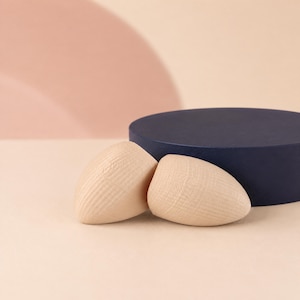 Puede incluir: Dos objetos de madera de color claro y superficie lisa, con forma redondeada similar a una gota y textura de veta natural visible, situados frente a un disco circular de color azul marino oscuro sobre una superficie plana y clara.