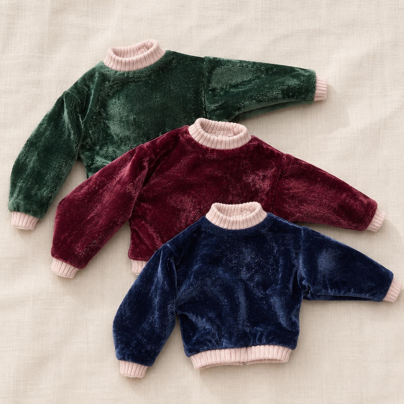 Op de afbeelding: Drie truien met lange mouwen in donkergroen, diepbordeaux en marineblauw. Elke trui heeft een zachte, fluweelachtige textuur, een geribbelde opstaande kraag en bijpassende geribbelde boorden aan de mouwen en zoom in lichtbeige.