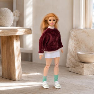 Op de afbeelding: Een pop met lang gemberkleurig haar draagt een donkerbordeauxrode fluwelen top met lange mouwen en een lichtroze opstaande kraag, een korte witte rok, lichtgroene enkelsokken en witte sneakers. De pop heeft scharnierende knie- en ellebooggewrichten.