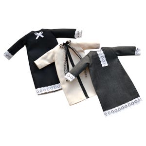 K&ouml;nnte beinhalten: Drei lang&auml;rmelige Puppenkleider in Schwarz, Creme und Anthrazit. Das schwarze Kleid hat eine wei&szlig;e Schleife und Spitzenbesatz. Das cremefarbene Kleid besitzt ein schwarzes Schleifenband und Spitzenb&uuml;ndchen. Das anthrazitfarbene Kleid hat eine Spitzenblende.