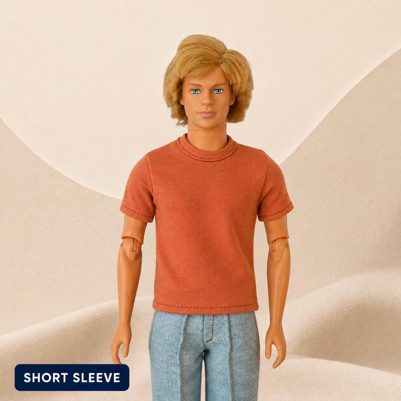 Puede incluir: Una mu&ntilde;eca con cabello rubio y ojos azules que viste una camiseta de manga corta de color &oacute;xido s&oacute;lido y pantalones de mezclilla azul claro. La mu&ntilde;eca tiene brazos articulados. Se observa una etiqueta rectangular azul oscuro con el texto SHORT SLEEVE.