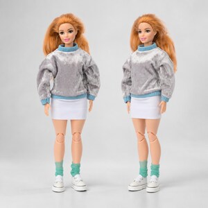 Op de afbeelding: Twee identieke poppen met lang oranje haar, gekleed in grijze fluwelen truien met lange mouwen met lichtblauwe geribbelde kragen en boorden, gecombineerd met effen witte minirokken, lichtblauwe sokken en witte sneakers.