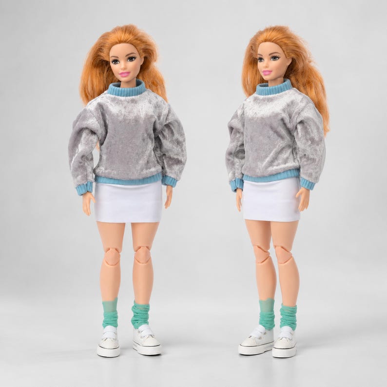 Op de afbeelding: Twee identieke poppen met lang oranje haar, gekleed in grijze fluwelen truien met lange mouwen met lichtblauwe geribbelde kragen en boorden, gecombineerd met effen witte minirokken, lichtblauwe sokken en witte sneakers.
