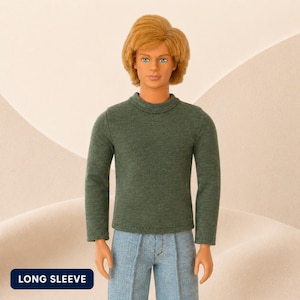 Puede incluir: Una mu&ntilde;eca con cabello rubio y ojos azules que viste un su&eacute;ter de manga larga de color verde oscuro y pantalones de mezclilla azul claro. La mu&ntilde;eca tiene una expresi&oacute;n facial neutral. Se ve una etiqueta rectangular azul oscuro con el texto LONG SLEEVE.