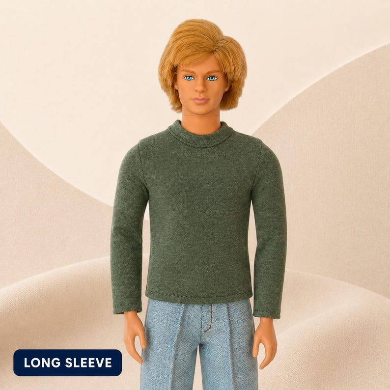 Puede incluir: Una mu&ntilde;eca con cabello rubio y ojos azules que viste un su&eacute;ter de manga larga de color verde oscuro y pantalones de mezclilla azul claro. La mu&ntilde;eca tiene una expresi&oacute;n facial neutral. Se ve una etiqueta rectangular azul oscuro con el texto LONG SLEEVE.