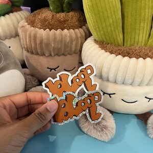 May include: A hand holds a sticker with the word "bloop" in orange, holographic lettering. The sticker is in front of several plush cactus toys with smiling faces and brown, fuzzy "soil" in their pots. The background is a light blue surface.