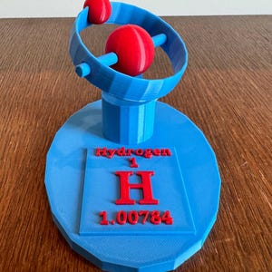 Puede incluir: Un modelo de átomo de hidrógeno impreso en 3D. El modelo es azul y rojo y presenta un electrón girando alrededor de un núcleo. La base del modelo incluye el símbolo del elemento "H" y el número atómico "1".