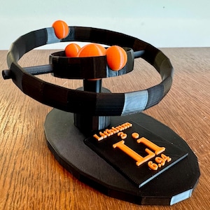 Puede incluir: Un modelo de átomo de litio impreso en 3D. El modelo presenta un anillo negro con tres esferas naranjas que representan electrones orbitando un núcleo negro. La base del modelo incluye un cuadrado negro con el texto "Lithium 3 Li 6.94" en naranja.