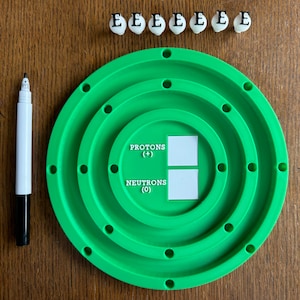 May include: A green plastic model of an atom with seven white letter 'E's on a wooden surface. The model has three rings with holes for the electrons. The center of the model has two white boxes labeled 'Protons (+) and Neutrons (0)'