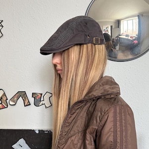 Könnte beinhalten: Braune Flatcap mit strukturiertem Design und kleiner Schnalle. Die Kappe wird mit einer braunen Jacke mit hohem Kragen und Knöpfen getragen. Im Hintergrund ist ein runder Spiegel zu sehen.