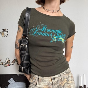 May include: A dark olive green short-sleeve t-shirt featuring the text "Romantic Summer 1953" in blue script with a floral graphic, paired with camouflage print trousers featuring a pattern of brown and tan leaves and tree branches.
