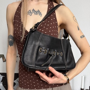 Könnte beinhalten: Schwarze Leder-Umhängetasche mit weißen Nähten und Kordelzugverschluss. Die Tasche hat einen kurzen Henkel und goldfarbene Beschläge. Die Tasche wird vor einer Person gehalten, die ein braunes Polka-Dot-Oberteil und einen karierten Rock trägt.