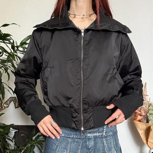 May include: Black bomber jacket with a wide collar and ribbed cuffs. The jacket is zipped up and paired with a denim mini skirt. The outfit is styled with a silver necklace and ring.