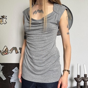 May include: A gray, sleeveless top with a draped cowl neckline. The top has a fitted silhouette and shoulder detailing. The model is wearing dark blue denim pants. The background includes wall art and a small side table with candles.