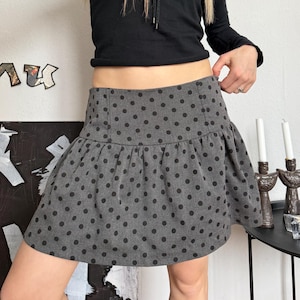 Könnte beinhalten: Ein schwarzes Langarm-Top mit Schnurzug-Ausschnitt und ein grauer Minirock. Der Rock hat ein schwarzes Polka-Dot-Muster und einen Rüschensaum. Der Rock ist etwa 30 cm lang.