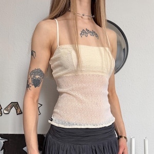 Könnte beinhalten: Ein cremefarbenes Spaghettitop mit einem durchsichtigen, strukturierten Overlay. Das Top wird mit einem dunkelgrauen, plissierten Rock kombiniert. Das Outfit wird durch eine silberne Halskette und ein Armband ergänzt.