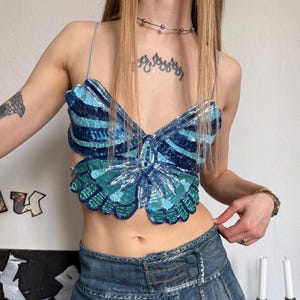 May include: A blue sequined butterfly top with thin straps and a denim mini skirt. The top features shades of blue and green sequins. The skirt is a classic denim wash with pleats.