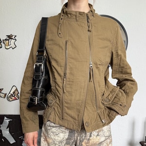 Op de afbeelding: Een bruine jas met meerdere ritsen en een hoge kraag. De jas heeft een getailleerd silhouet en heeft een klein zakje op de borst. De persoon draagt ook een camouflagebroek en een zwarte leren schoudertas.