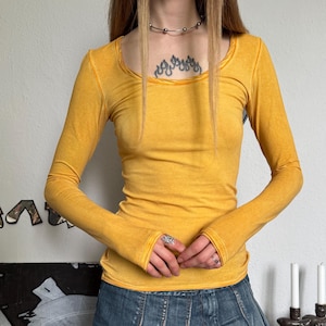 Könnte beinhalten: Senfgelbes Langarm-Top mit Rundhalsausschnitt und figurbetonter Silhouette. Das Top wird mit einem Jeans-Minirock kombiniert. Der Rock hat ein plissiertes Design und eine verwaschene Optik. Das Outfit ist lässig und stilvoll.